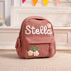 Rosa Corduroy Rucksack mit eingesticktem Namen Stella und floralen Details auf einem hellen Teppich.