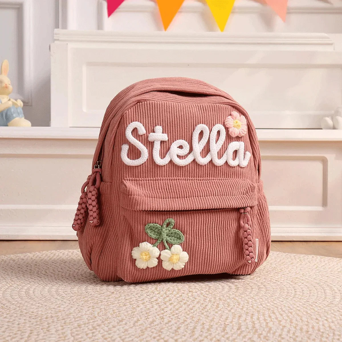 Rosa Corduroy Rucksack mit eingesticktem Namen Stella und floralen Details auf einem hellen Teppich.