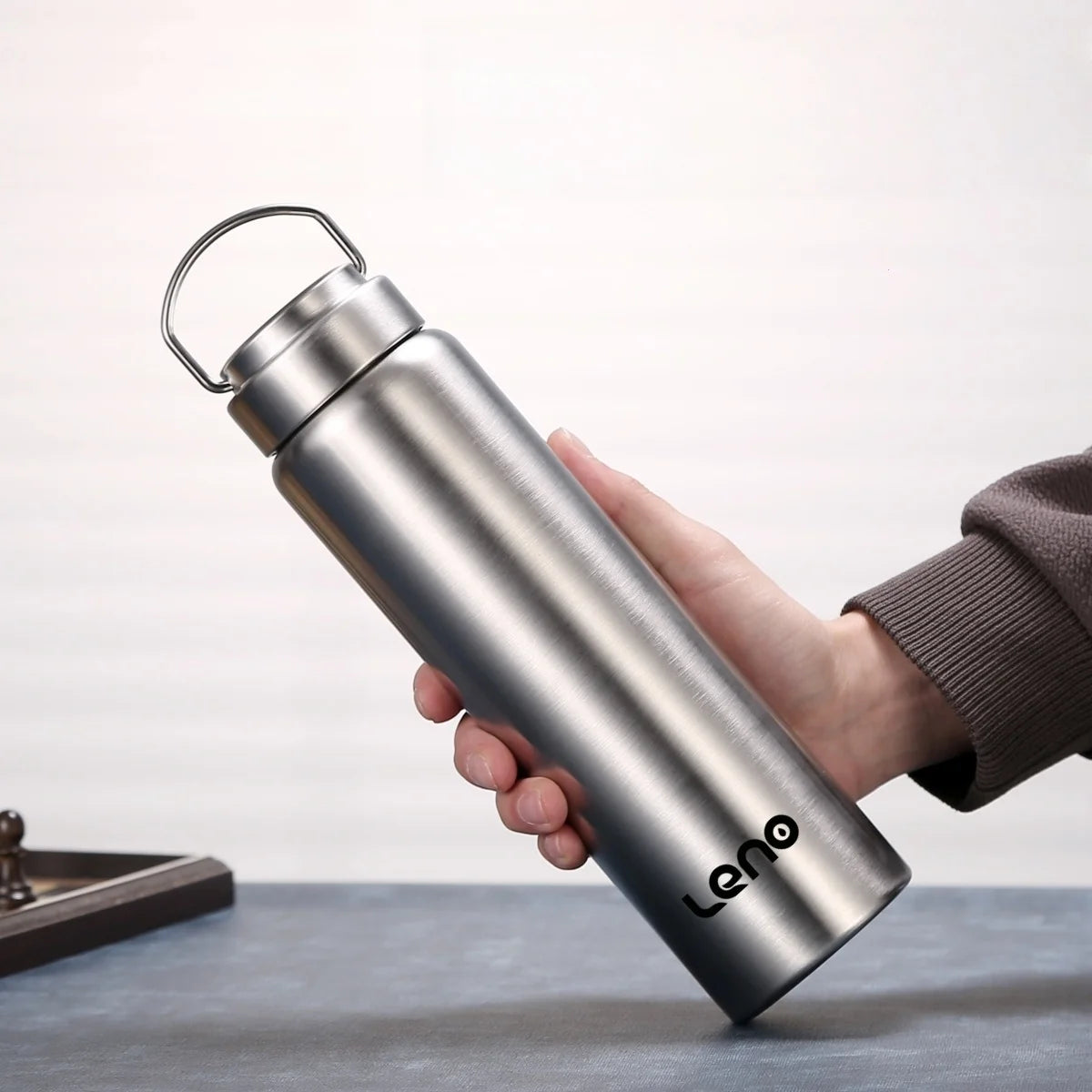 A hand holds a silver stainless steel sports water bottle with a hinged lid against a light background.