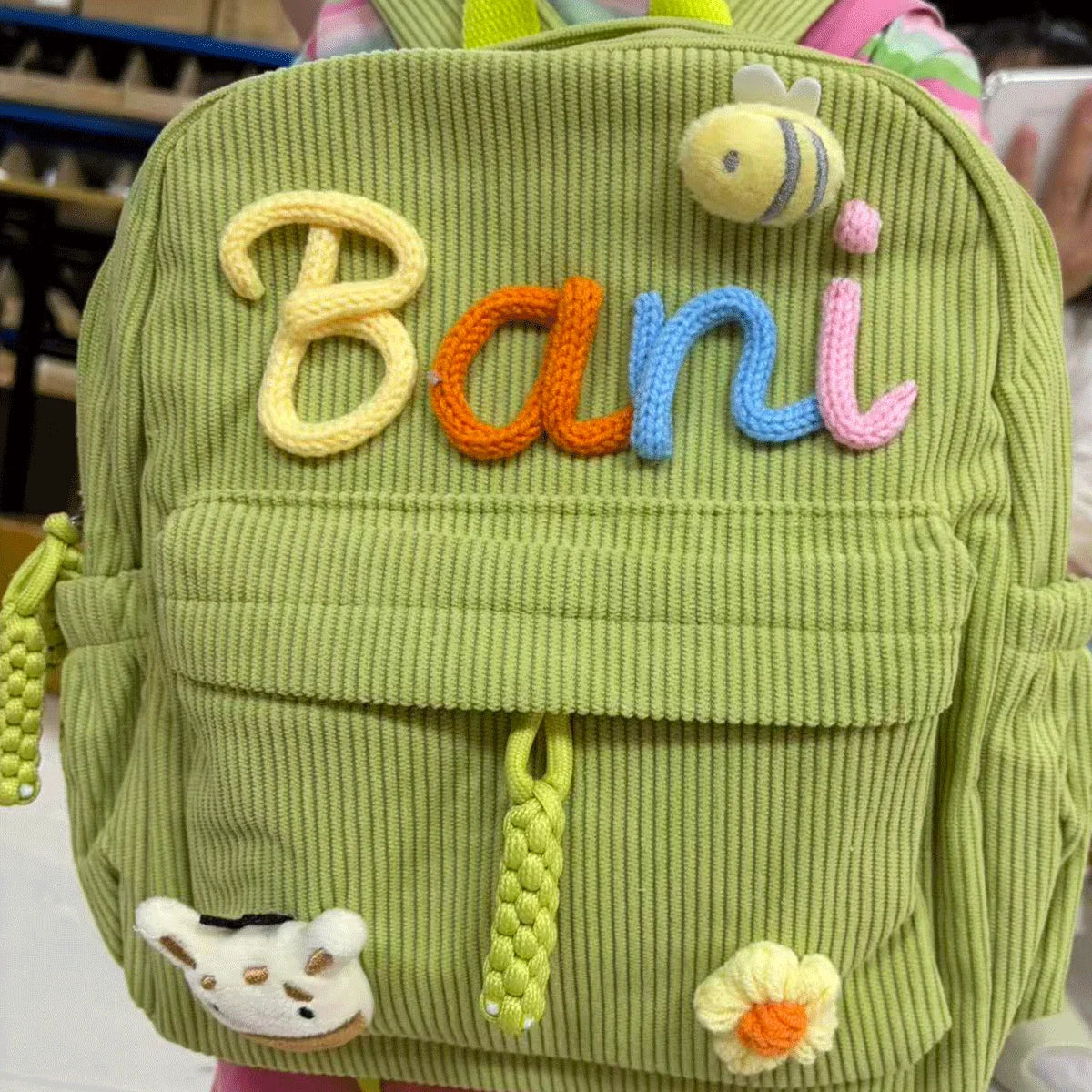 Helles grün-cordhaltiges Rucksack mit dem Namen Bani in orange- und blauen Buchstaben und einer gelben Biene.