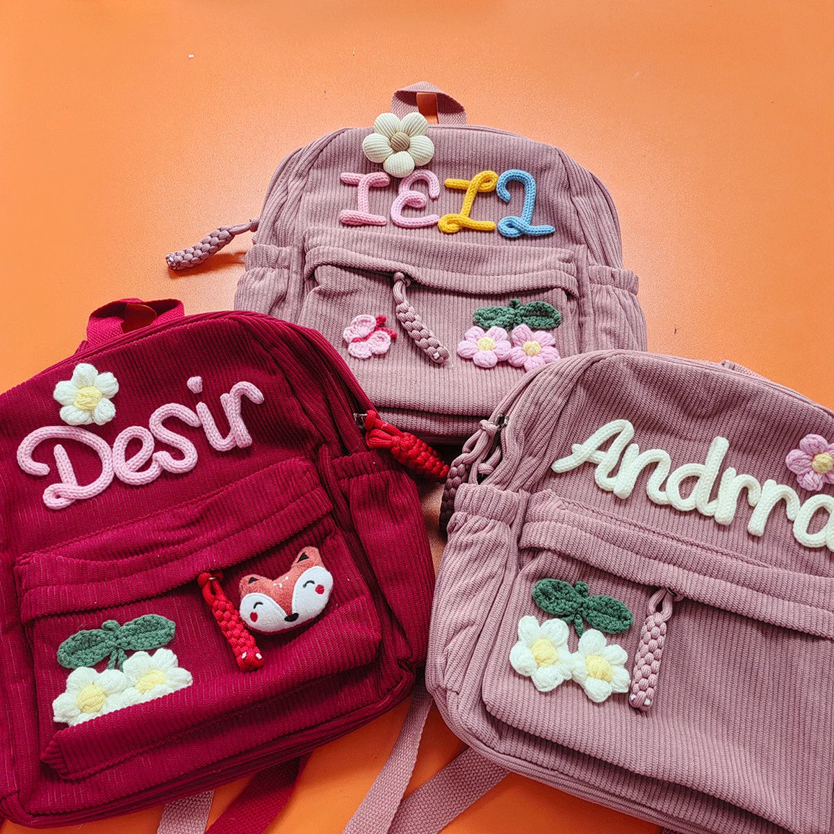 Eine Kollektion von rosa und kastanienbraunen Cord-Rucksäcken mit gestickten Buchstaben, Blumen und Tieren.