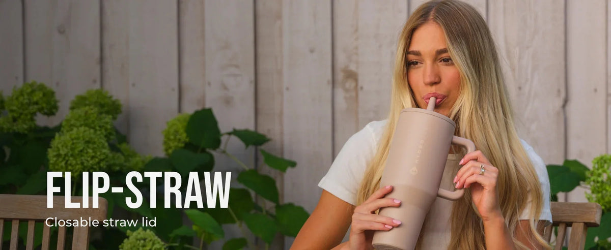 A blonde woman holds a beige insulated water bottle with a flip straw against a wooden background.