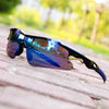 A pair of blue and purple sport sunglasses resting on a wooden surface with a blurred green outdoor background.