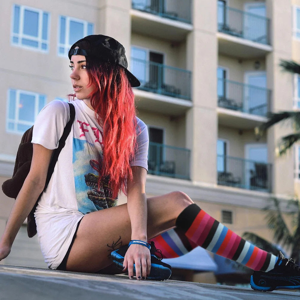 A woman with long red hair wearing a white shirt and black hat is sitting outdoors, modeling medical grade compression socks.