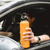 An orange double-wall insulated stainless steel thermos is being held by a person inside a car.
