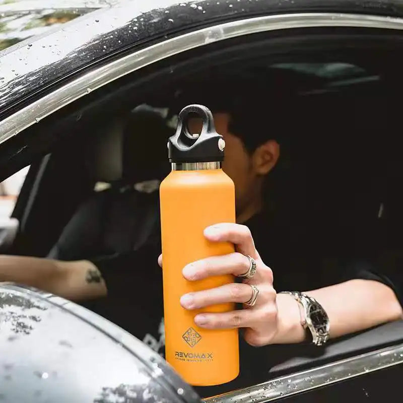 An orange double-wall insulated stainless steel thermos is being held by a person inside a car.