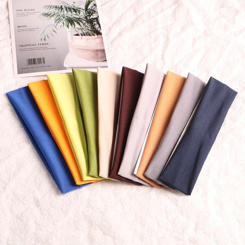 A collection of multi-use elastic headbands in various shades of green, yellow, beige, brown, and blue are displayed on a lig