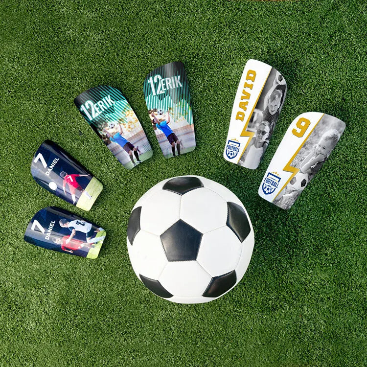 A collection of nine soccer shin guards featuring different player names and numbers on a green turf background.