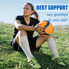 A woman wearing compression socks sits on grass holding an orange exercise ball.