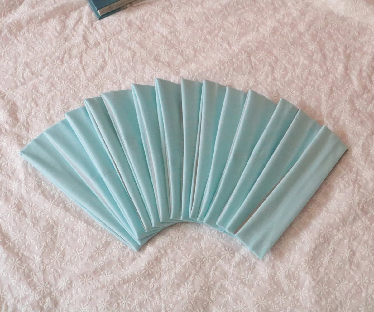 A fan of light blue satin elastic headbands arranged in a pleated fan shape on a pink textured surface.