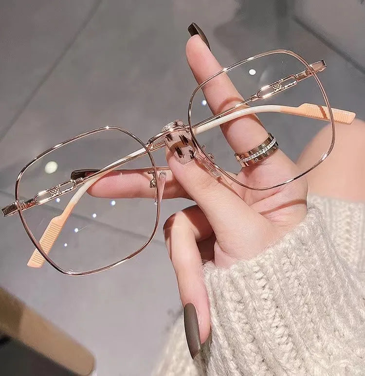 A hand holds a pair of thin, rose gold anti-blue light reading glasses with light wood accents against a blurred background.