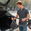 A man is using a black electric scooter for adults while the trunk of a silver car is open on a city street.