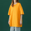 Eine Frau in einem leuchtend orangefarbenen, oversized Baumwoll-T-Shirt und hellgrauen Hosen vor einem dunkelgrünen Hintergru