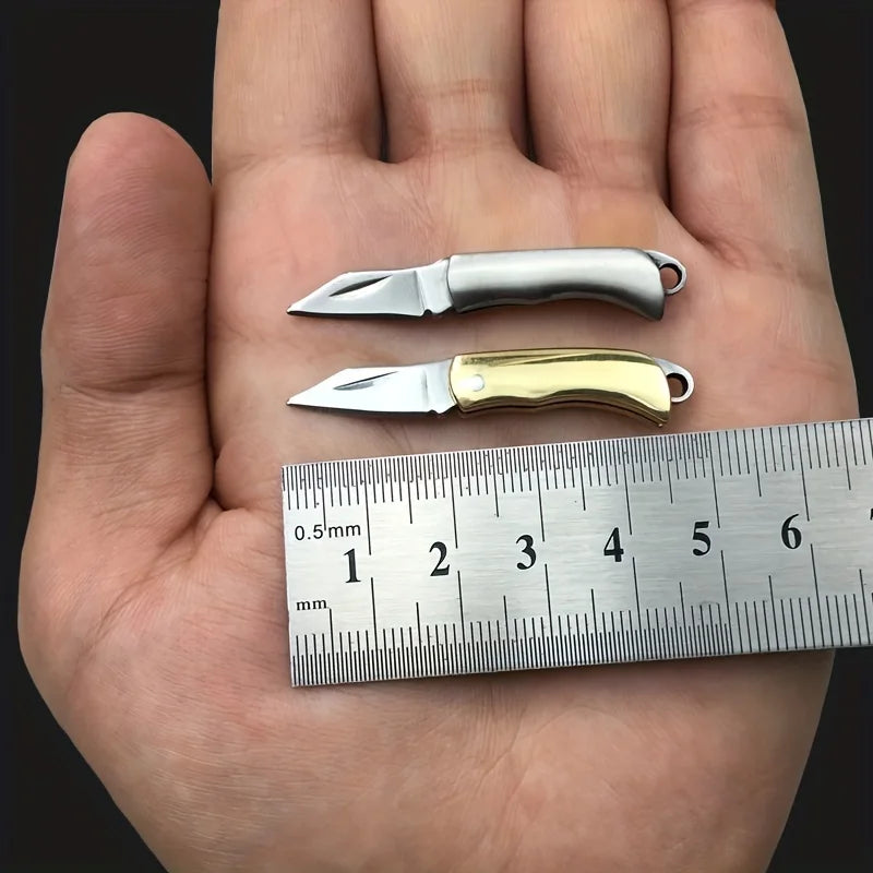 Una mano sostiene un cuchillo de llavero plegable plateado y uno dorado junto a una regla.