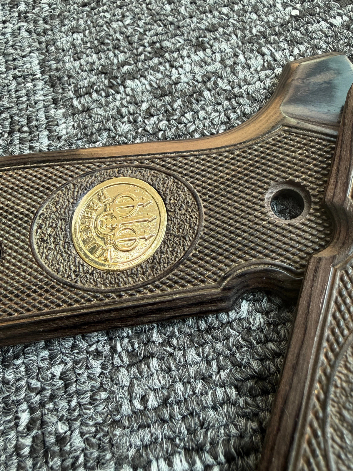 Close-up of a wooden firearm grip with diamond pattern grips and a brass medallion.