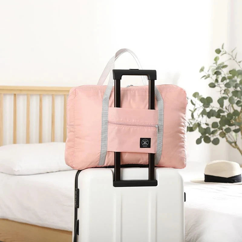 A pink foldable travel duffel bag with a black handle is placed on top of a white rolling suitcase in a bedroom setting.