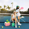 A woman in a white top and skirt holds a pink paddle and sits on a chair near a blue sports court with water bottles.