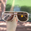 A hand holds a pair of photochromic square frame sunglasses with UV400 protection against a blurred outdoor background.