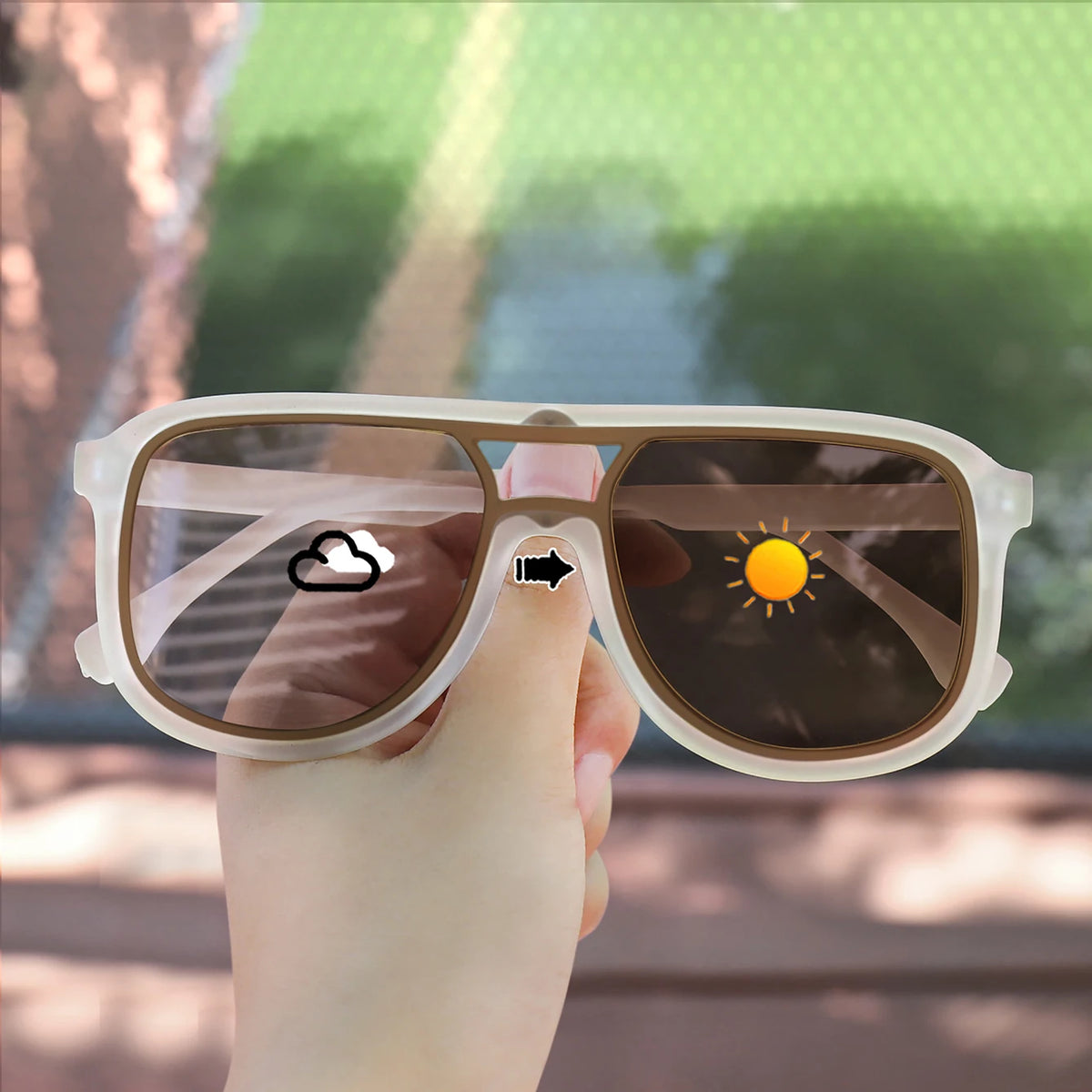 A hand holds a pair of photochromic square frame sunglasses with UV400 protection against a blurred outdoor background.