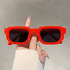 A hand holds a pair of bright orange rectangular sunglasses against a light gray background.