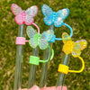 A collection of colorful silicone butterfly straw covers on clear straws against a green outdoor background.