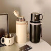 Une bouteille isotherme en acier inoxydable beige clair à côté d'une noire et d'une tasse argentée sur une surface blanche.
