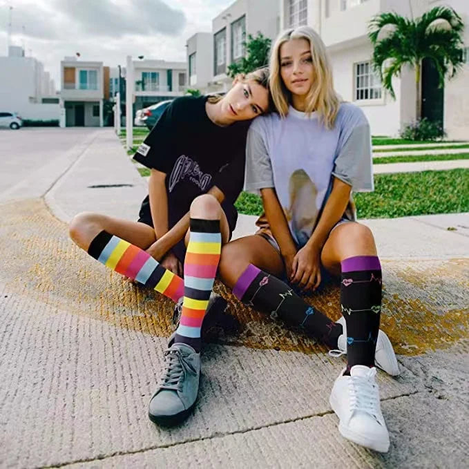 Two young women wearing colorful compression elastic socks sitting on a concrete sidewalk.