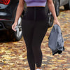 A woman wearing high waisted shaping leggings and a purple sports bra is walking outdoors on a leaf-covered path.