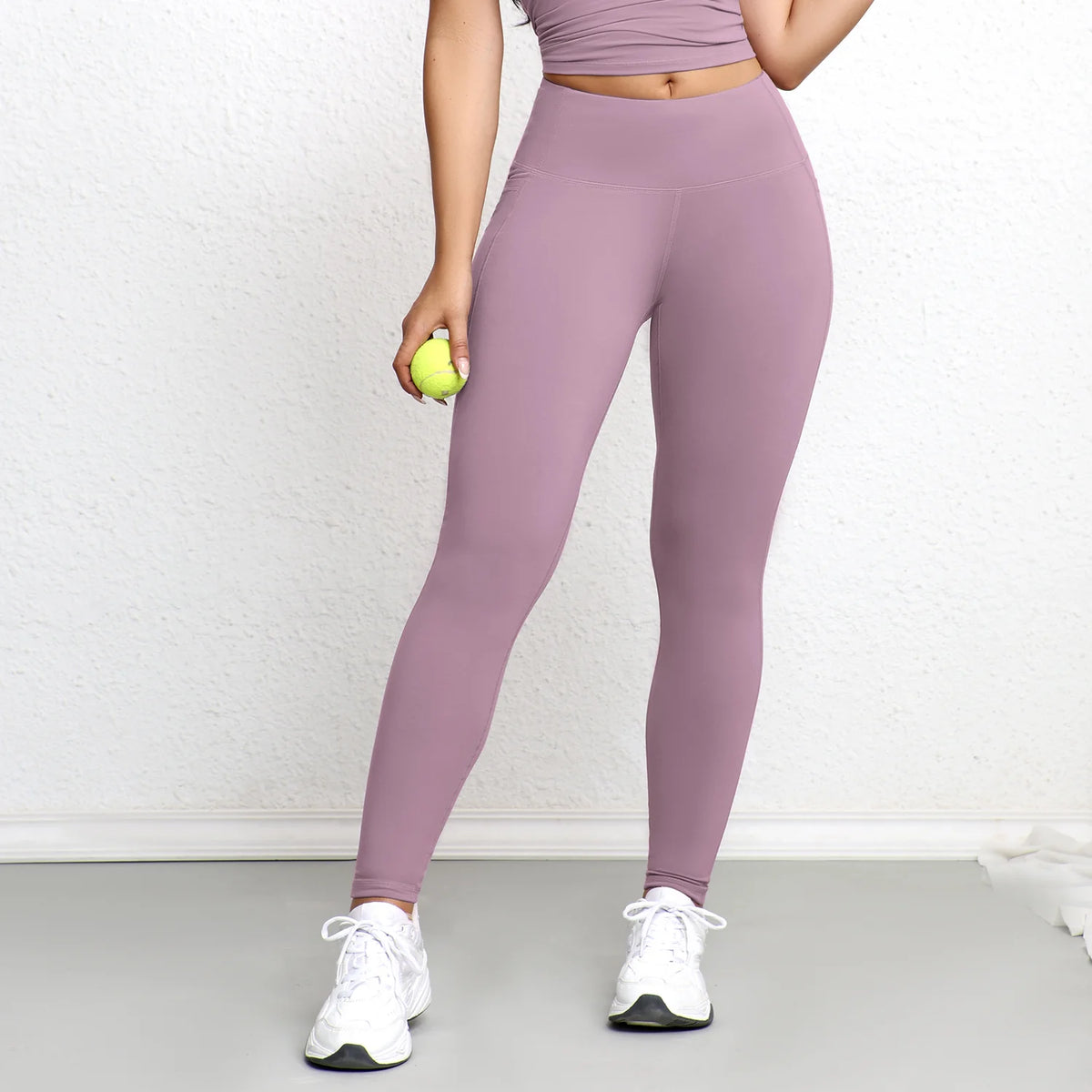 A woman wearing mauve high-waisted sports leggings and a purple top stands indoors.