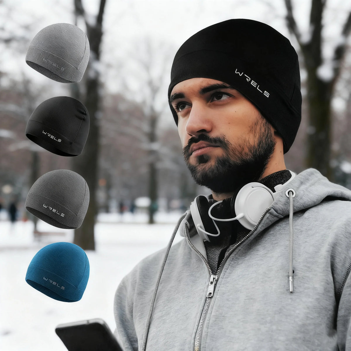 A man wearing a black beanie hat and headphones holds several knit hats in front of him against a snowy outdoor background.