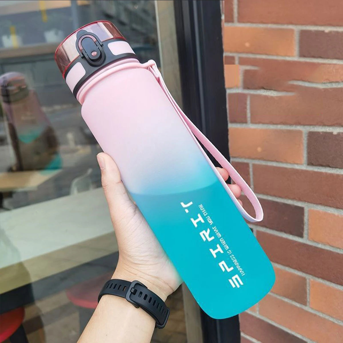 Eine Hand hält eine verlustige isolierte Wasserflasche mit rosa und blauen Akzenten vor einem Backstein-Hintergrund.