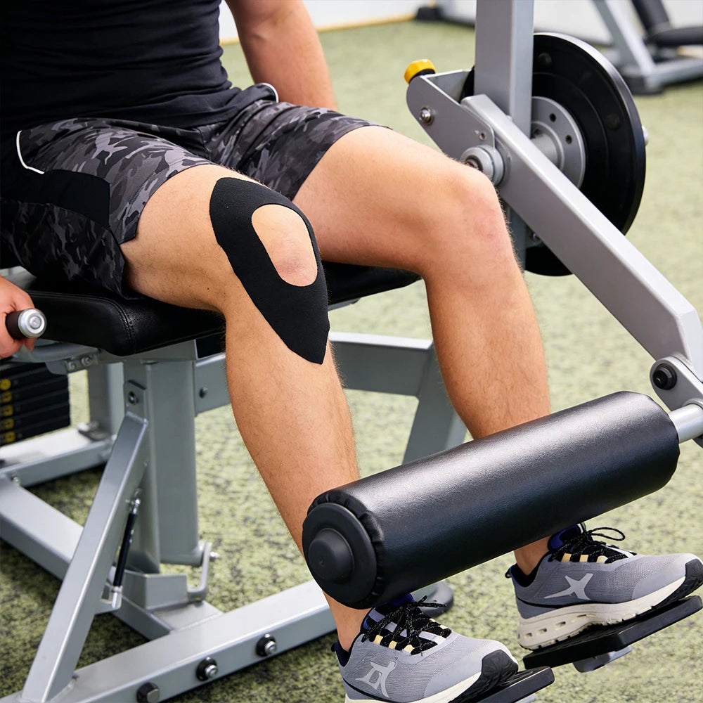 Eine Person verwendet Kinesiologie-Tapes am Knie beim Training auf einer Fitnessmaschine.