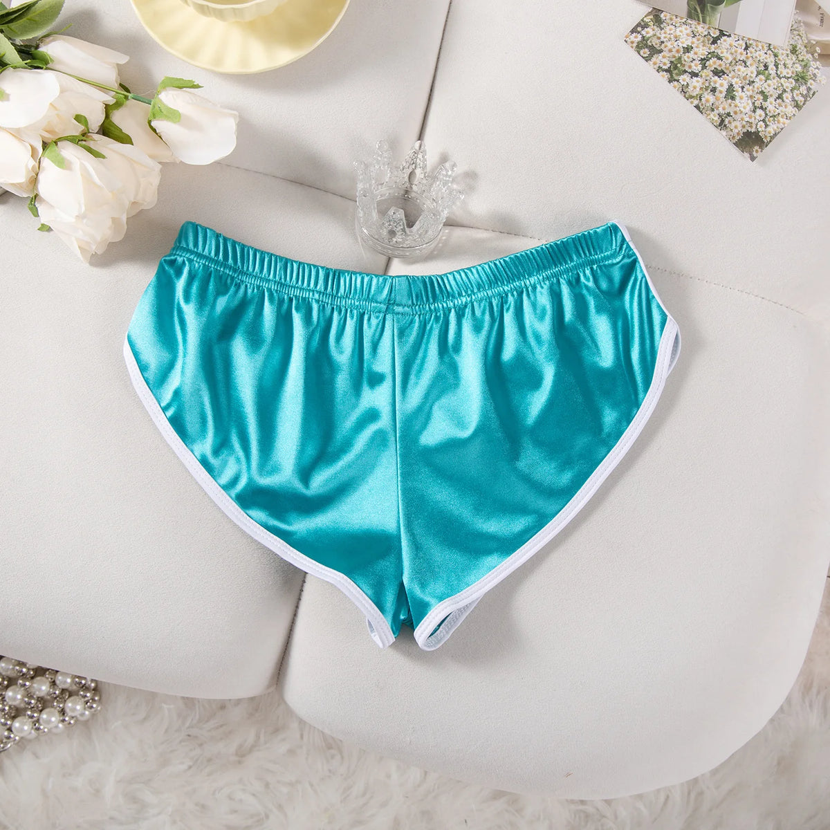 Ein Paar tealblauer Satin-Trainingsshorts mit floralen Akzenten auf einer weißen Oberfläche.