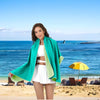 A woman wearing a bright teal microfiber quick-dry beach towel stands on a sandy beach with the ocean in the background.
