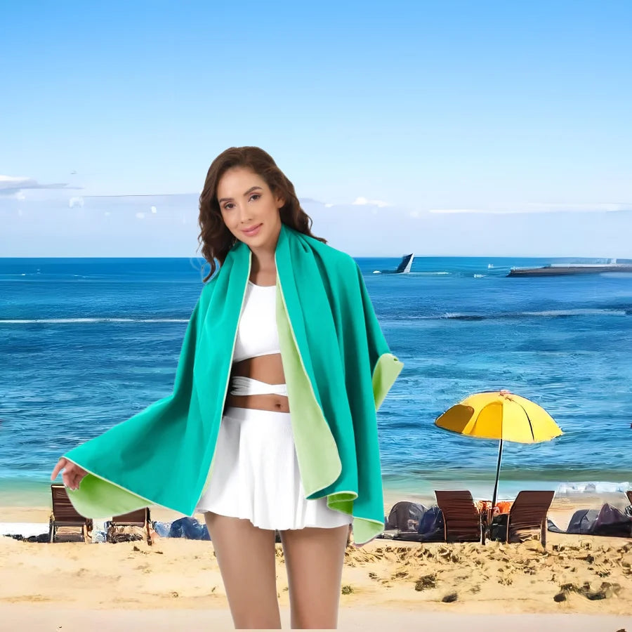 A woman wearing a bright teal microfiber quick-dry beach towel stands on a sandy beach with the ocean in the background.