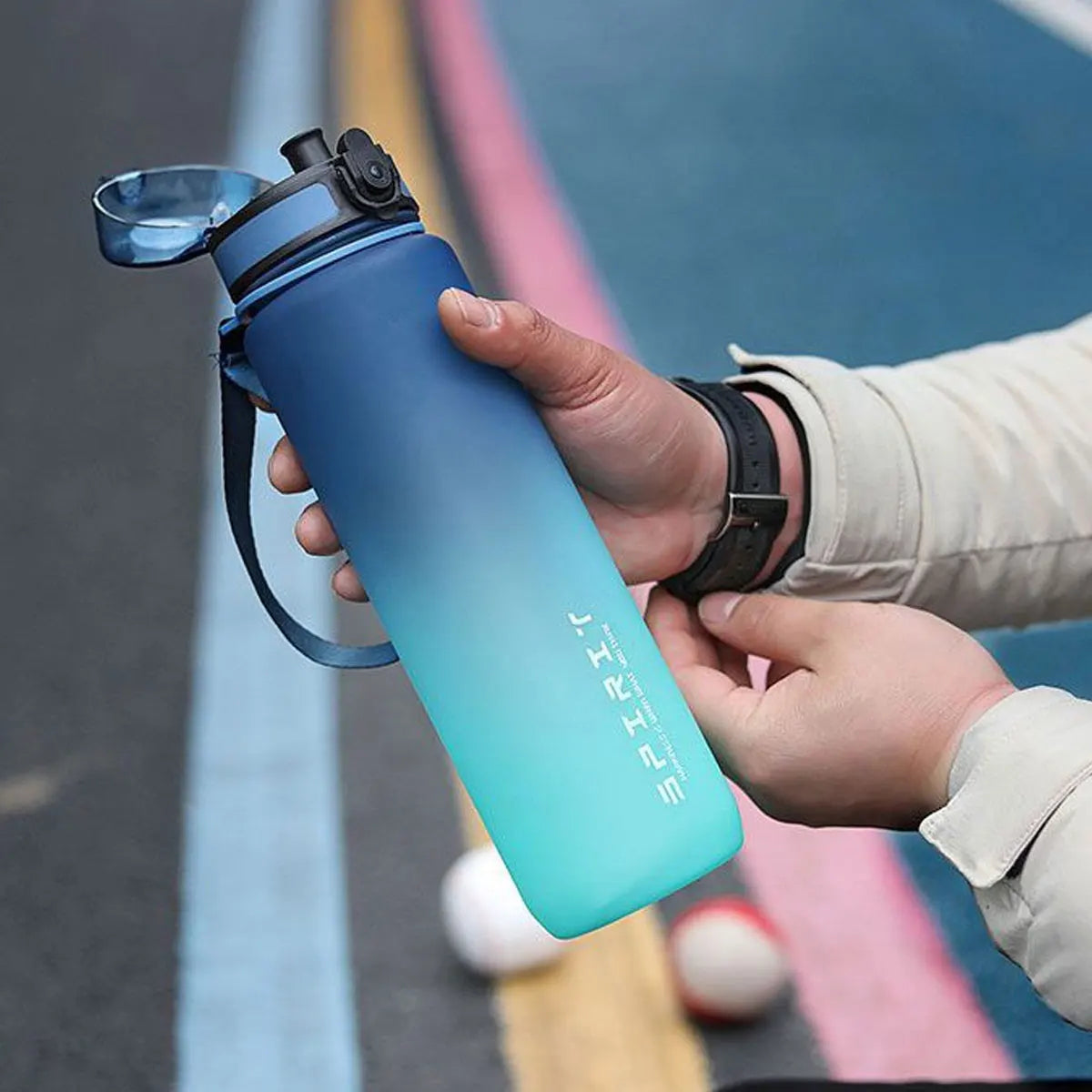 Eine Hand hält eine gradienten-isolierte Wasserflasche in blau-türkis gegen einen unscharfen Außenhintergrund.