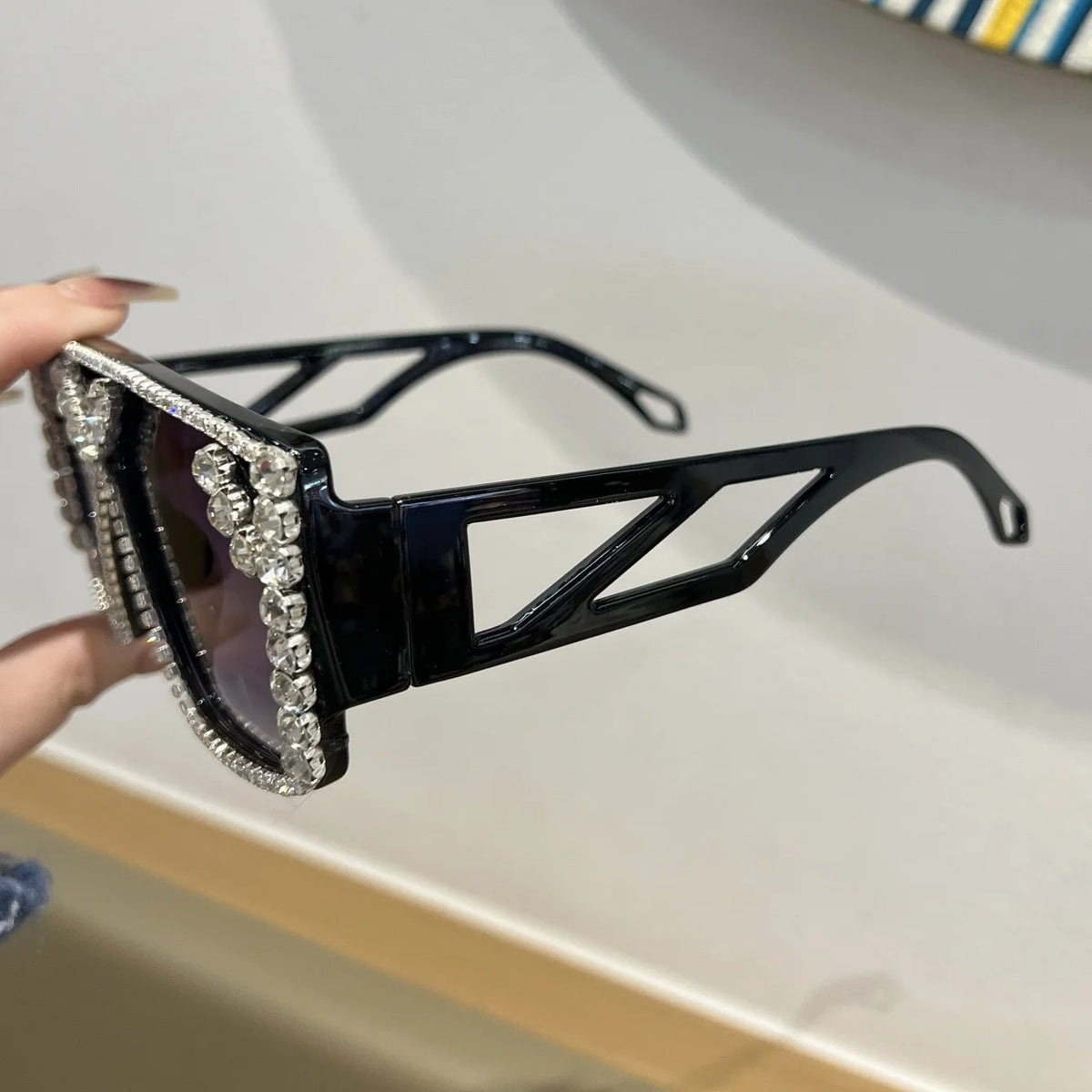 A pair of black geometric fashion sunglasses adorned with clear rhinestones, held by a hand.