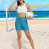 A woman wearing white athletic top and teal shorts stands on a beach with a tennis net in the background.