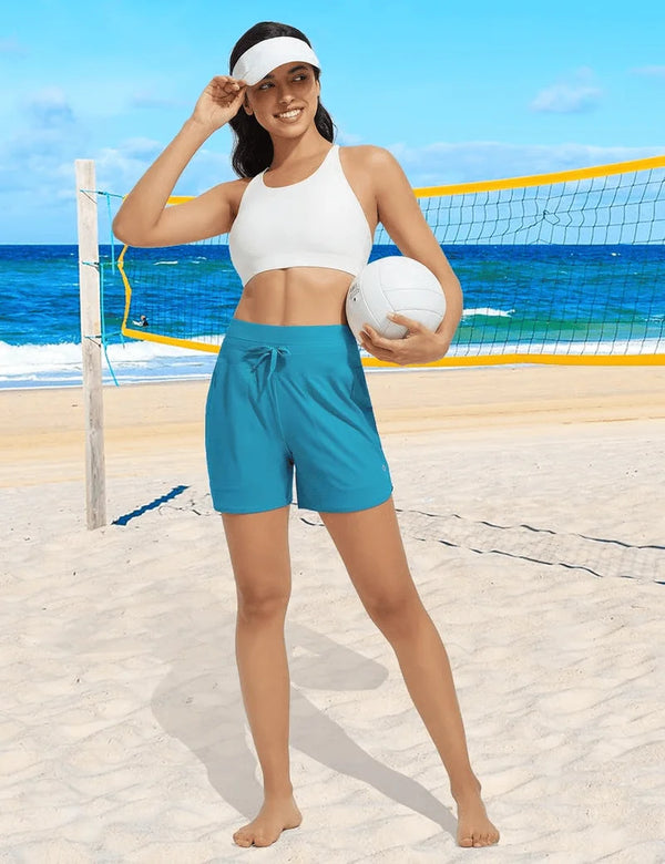 A woman wearing white athletic top and teal shorts stands on a beach with a tennis net in the background.