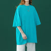 Eine Frau in einem tealfarbenen, oversized Baumwoll-T-Shirt und hellen Hosen vor einem dunkelgrünen Hintergrund.