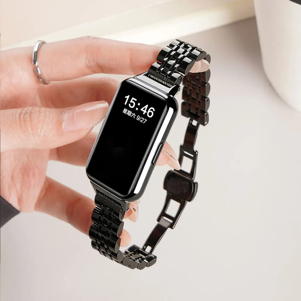 A hand is wearing a black smartwatch with a stainless steel mesh band displaying the time 15:46.