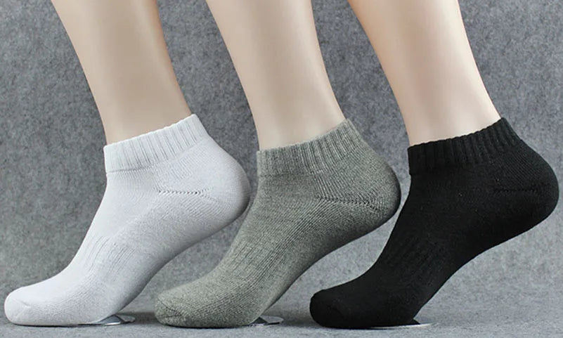 Un set de tres calcetines deportivos de algodón para el tobillo en blanco, gris y negro mostrados sobre una superficie.