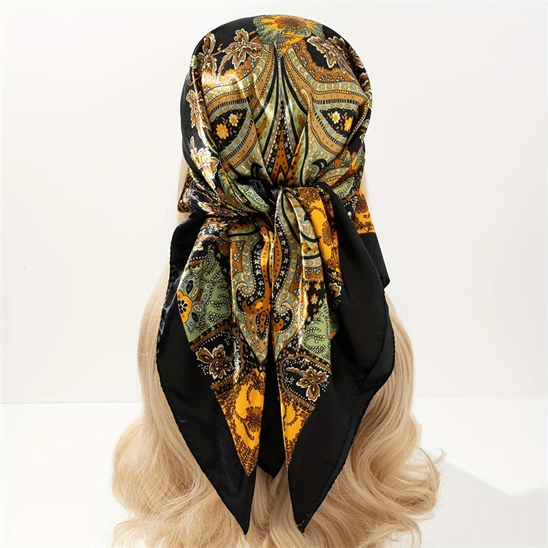 A black satin square scarf with an ornate gold and olive pattern tied in a bow, displayed on blonde hair.