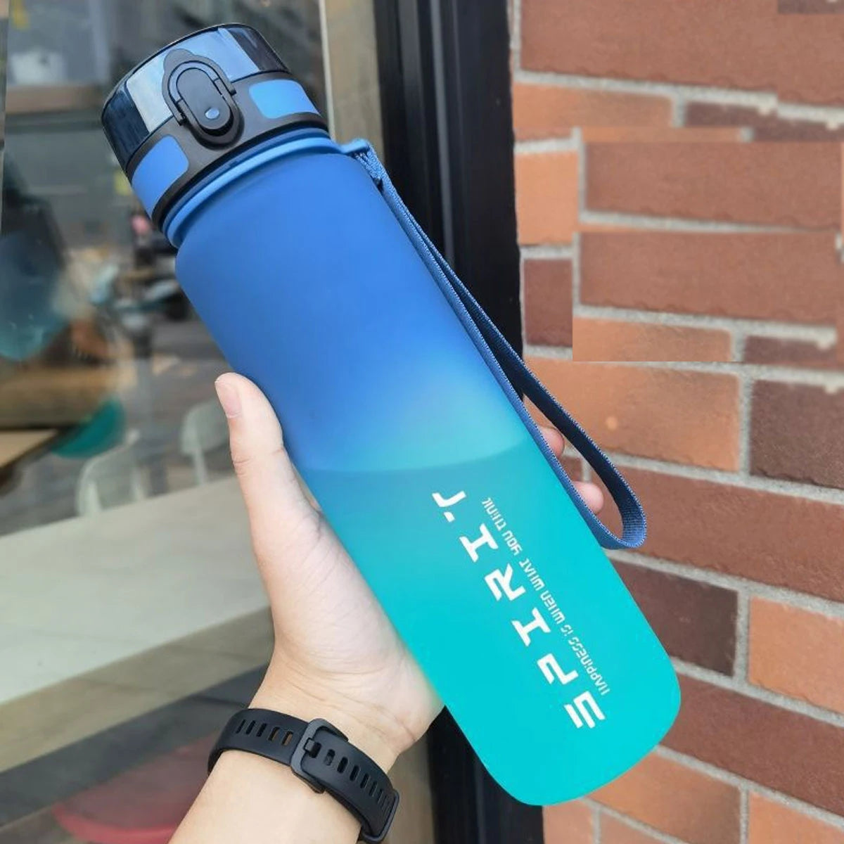 Eine Hand hält eine vergraffte isolierte Wasserflasche mit schwarzem Deckel und blau-grüner Farbe vor einer Ziegelwand.