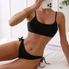 Eine Frau in einem schwarzen, hoch taillierten String-Bikini-Set posiert auf einem weißen Bett.