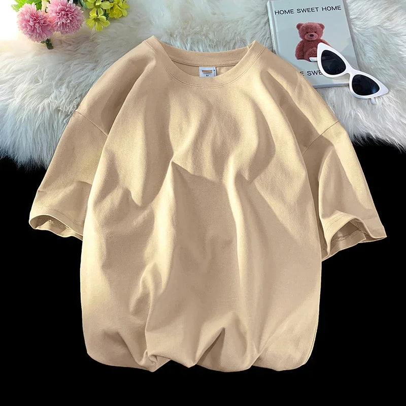Beige, überdimensioniertes Baumwoll-T-Shirt einer Frau auf weißem Plüsch neben einem Teddybär und Sonnenbrille.