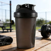 A matte black shaker bottle is sitting on a wooden surface next to a black towel and a dumbbell.