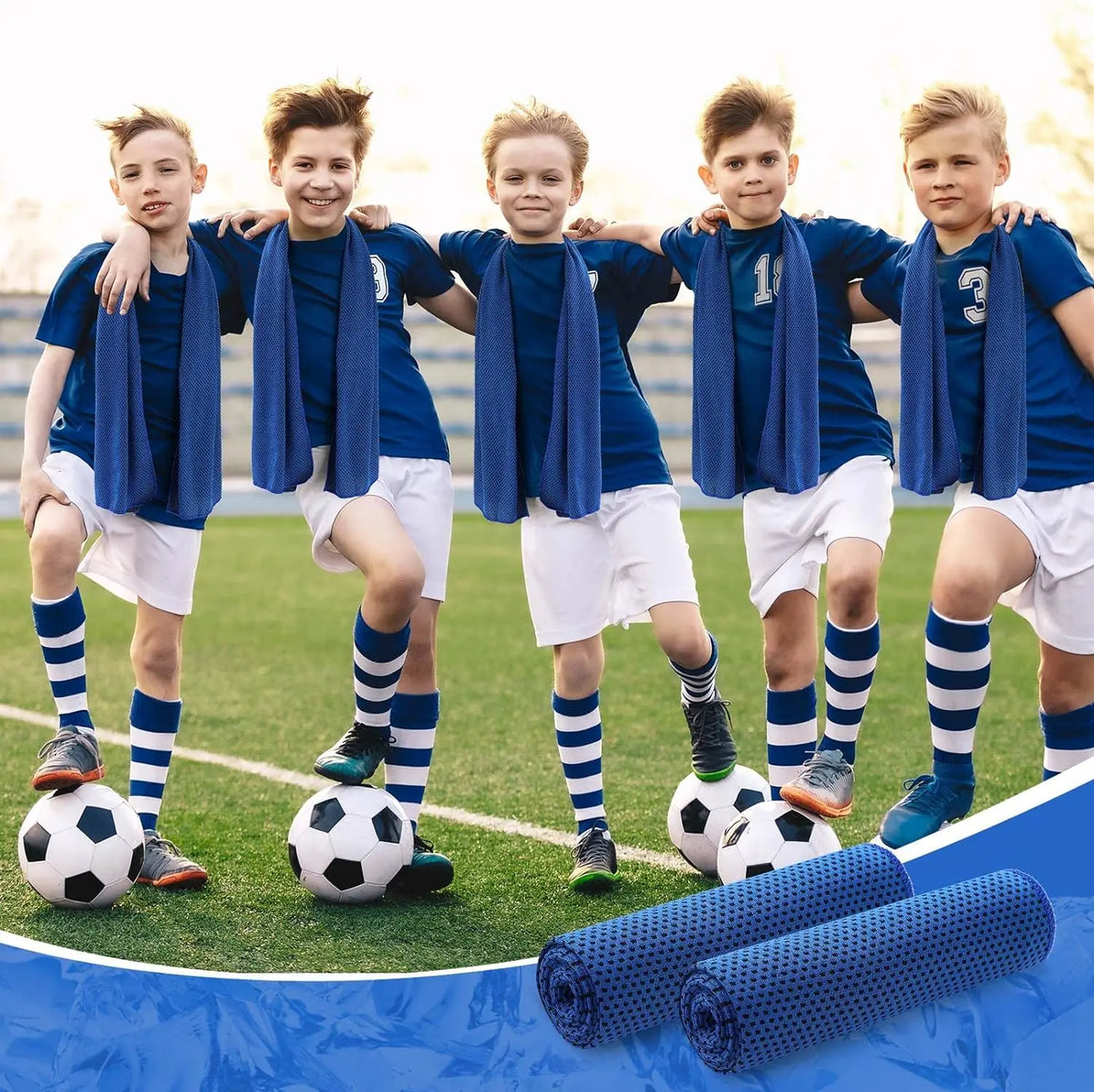 Five boys wearing blue shirts and white shorts standing on a green field with soccer balls and rolled microfiber towels.