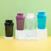 A set of four leakproof protein shaker bottles in purple, white, and blue are displayed on a light green background.
