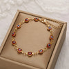 Gold charm bracelet with red and white evil eye beads displayed in a light brown box.